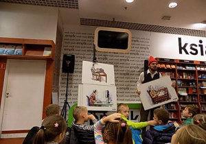 Mężczyzna w czapce krasnala prezentuje dzieciom trzy duże ilustracje.